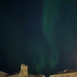 Bầu trời đêm với hiện tượng Bắc Cực Quang (Northern Lights) rực rỡ trên một thành phố, thể hiện hiệu ứng phơi sáng dài trong nhiếp ảnh điện thoại.