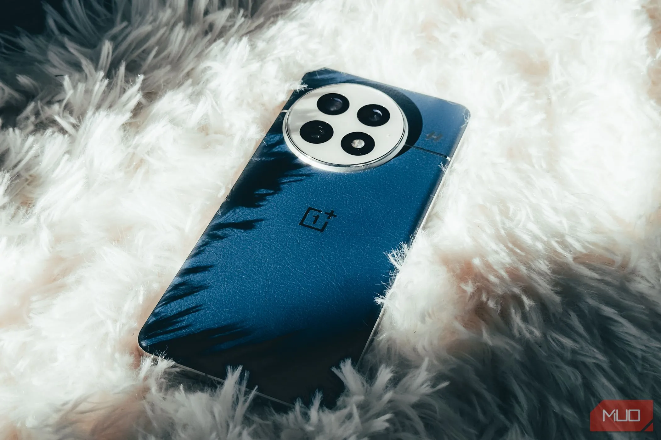Điện thoại OnePlus 13 với thiết kế mặt lưng da thuần chay màu xanh, đặt trên nền thảm trắng mềm mại, tôn lên vẻ cao cấp.