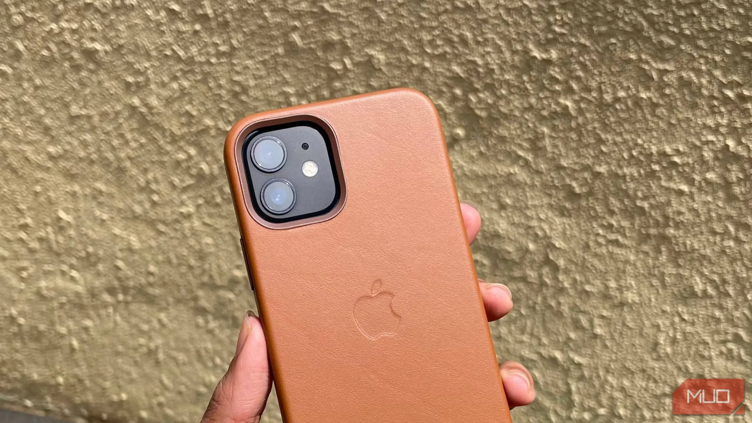 Một chiếc iPhone 12 màu vàng cát đặt trong ốp lưng da, được cầm trên tay, phía sau là bức tường. Ốp lưng có vẻ có gờ bảo vệ camera.