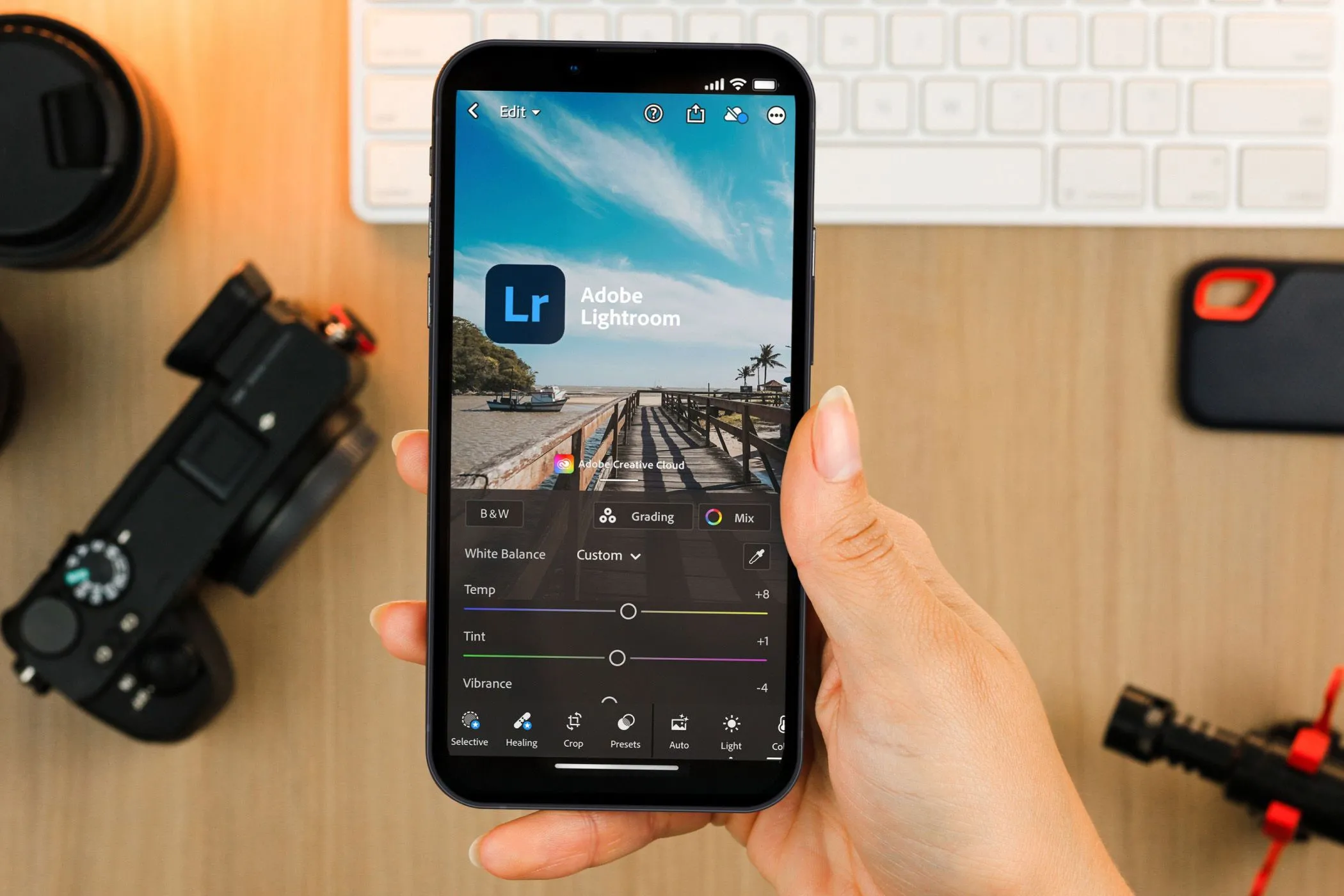 Người dùng đang sử dụng ứng dụng Lightroom Mobile trên điện thoại với thiết bị chụp ảnh chuyên nghiệp phía sau, nhấn mạnh tính năng chỉnh sửa ảnh chuyên sâu